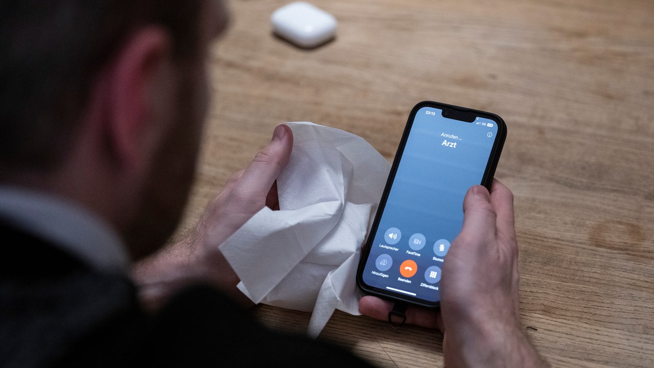 Ein Mann hält ein Taschentuch in einer Hand und ein Telefon mit dem Schriftzug «Arzt» in der Anderen. 