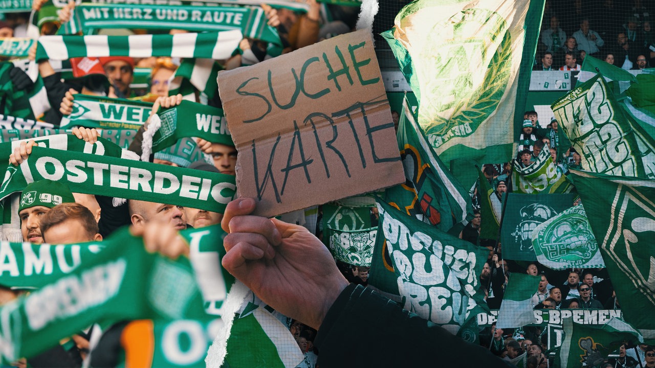 Eine Hand hält ein Schild hoch mit der Aufschrift "Suche Karte", dahinter sind Fotos von der Ostkurve mit Werder-Schal und -Fahnen.